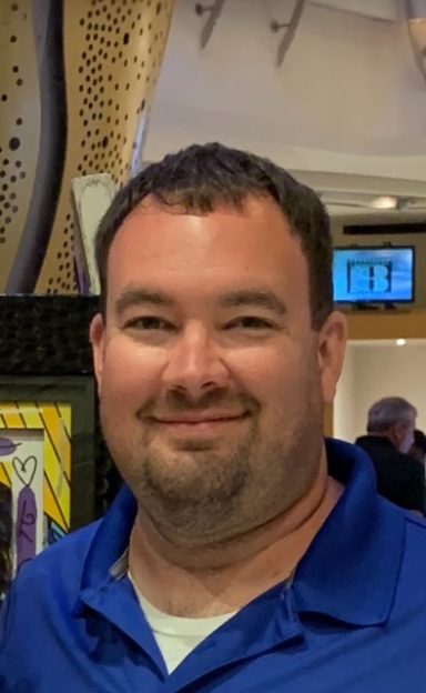 Smiling man with short brown hair, wearing a blue polo shirt in an indoor setting.